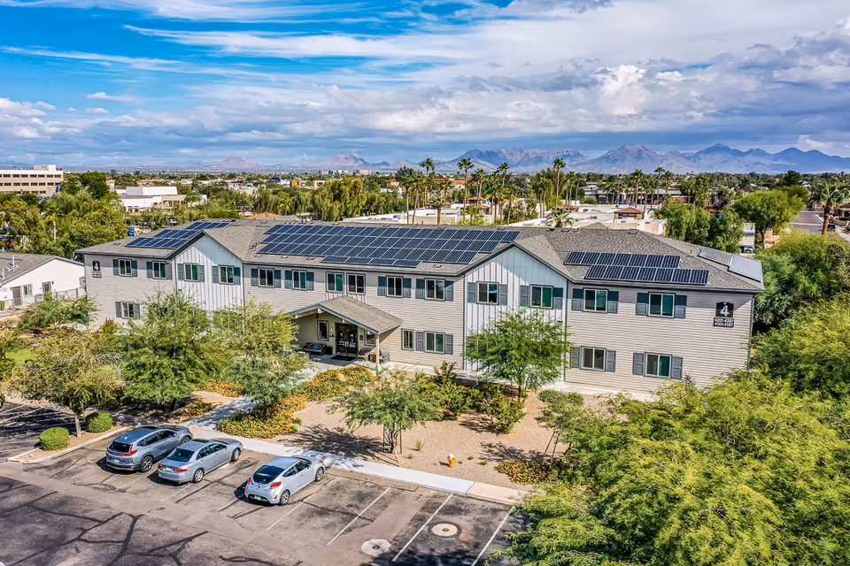 A two-story senior living building with solar panels on the roof, surrounded by trees and a parking lot with cars and mountains in the background.