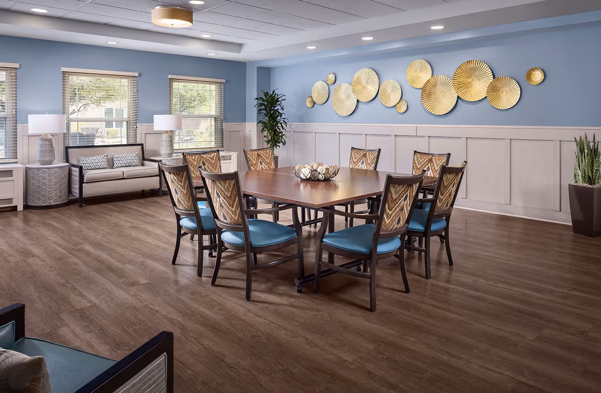 A spacious and well-lit dining room with a large square wooden table surrounded by eight chairs with patterned backs and blue seats. The room features light blue walls with white wainscoting, three windows with blinds, two table lamps on side tables, a small sofa with cushions, decorative golden wall art, and potted plants in the corners. The floor is wooden with a warm finish.