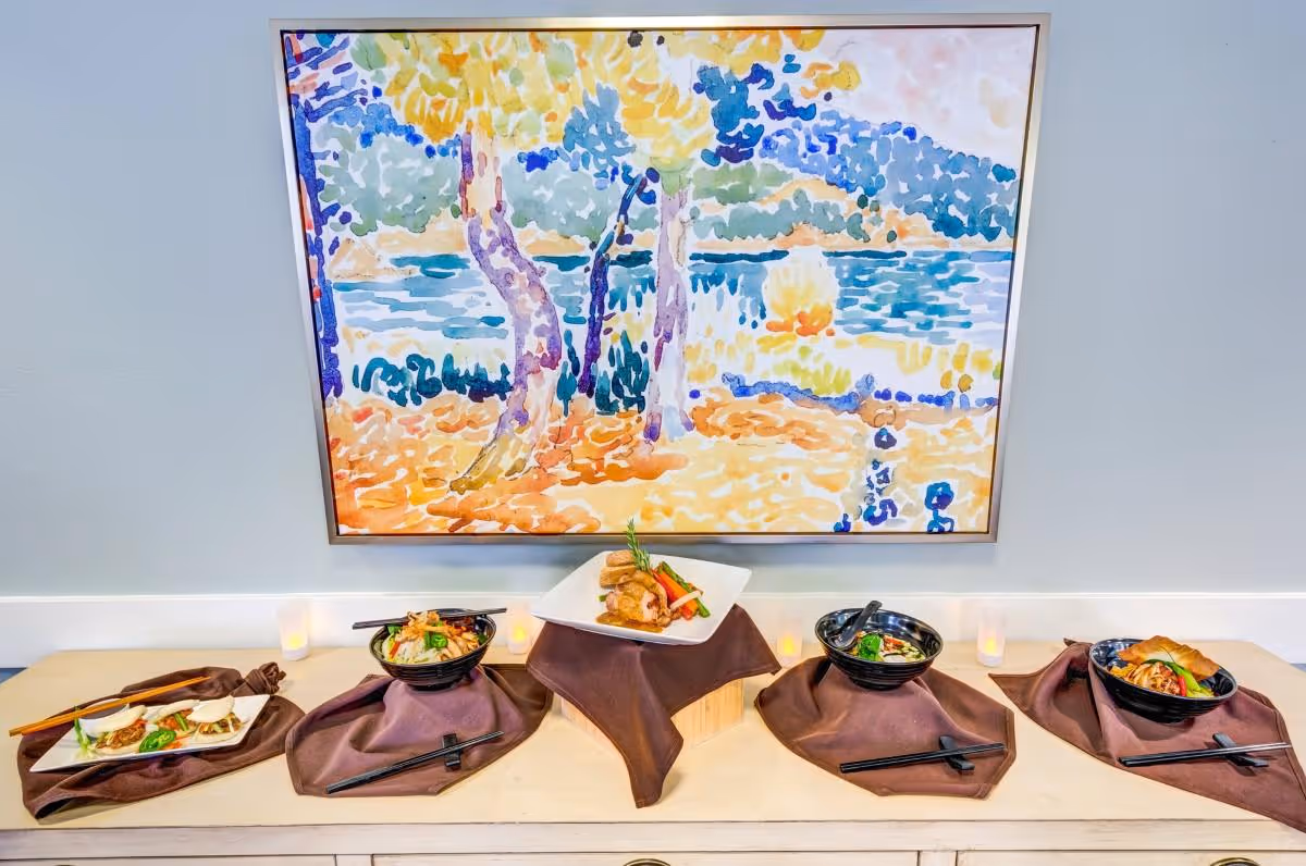 A display of various plated Asian-inspired dishes arranged on a wooden surface with brown cloth napkins underneath each plate. Behind the food is a colorful abstract painting depicting trees and a body of water.