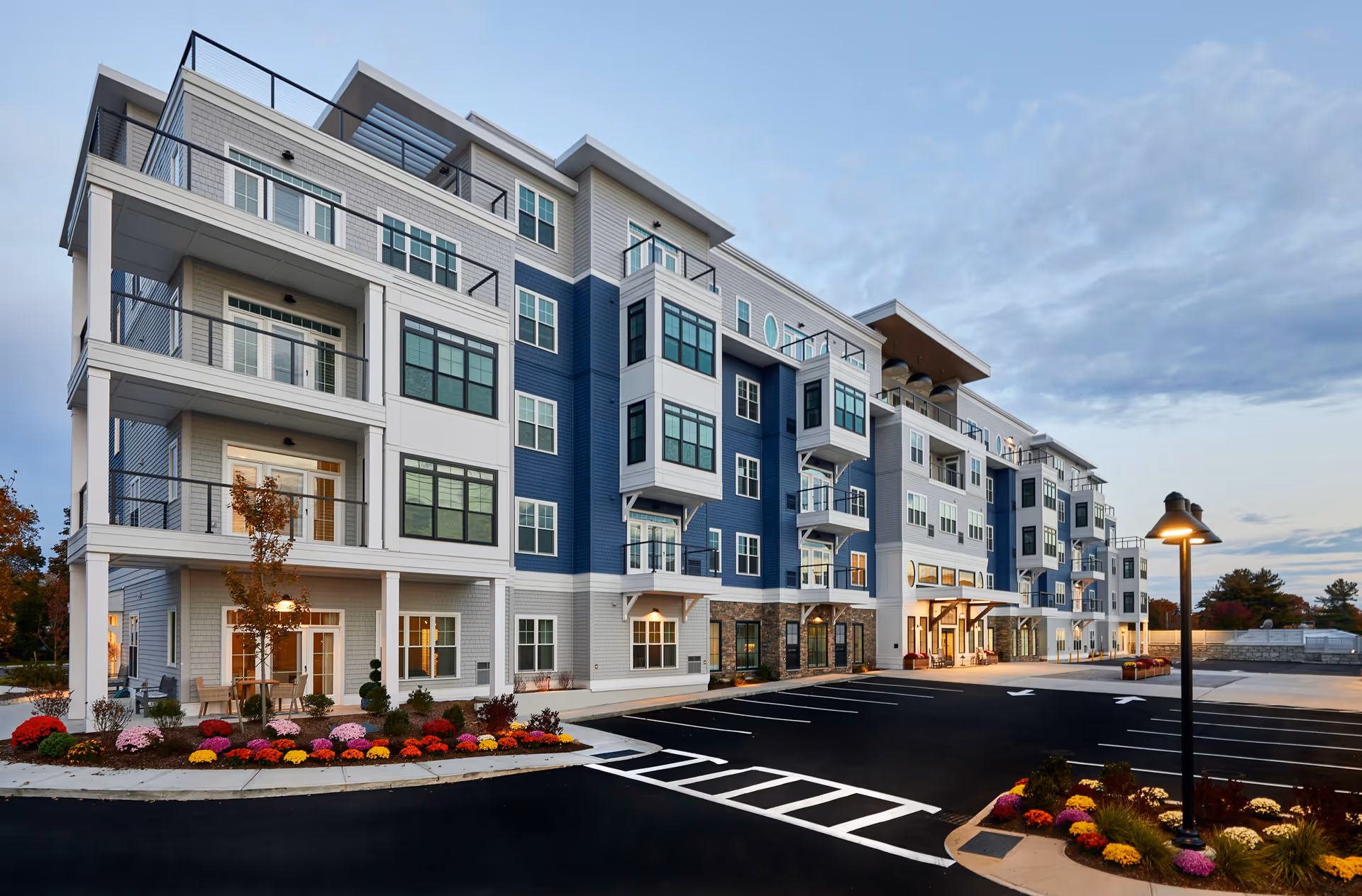 Four-story residential building with balconies, landscaped flowerbeds, and a paved parking lot at dusk.