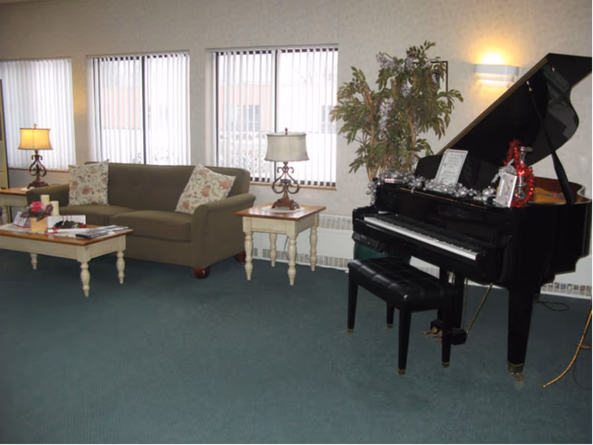 A cozy lounge with a sofa, coffee tables, lamps and a black grand piano in front of windows with vertical blinds.