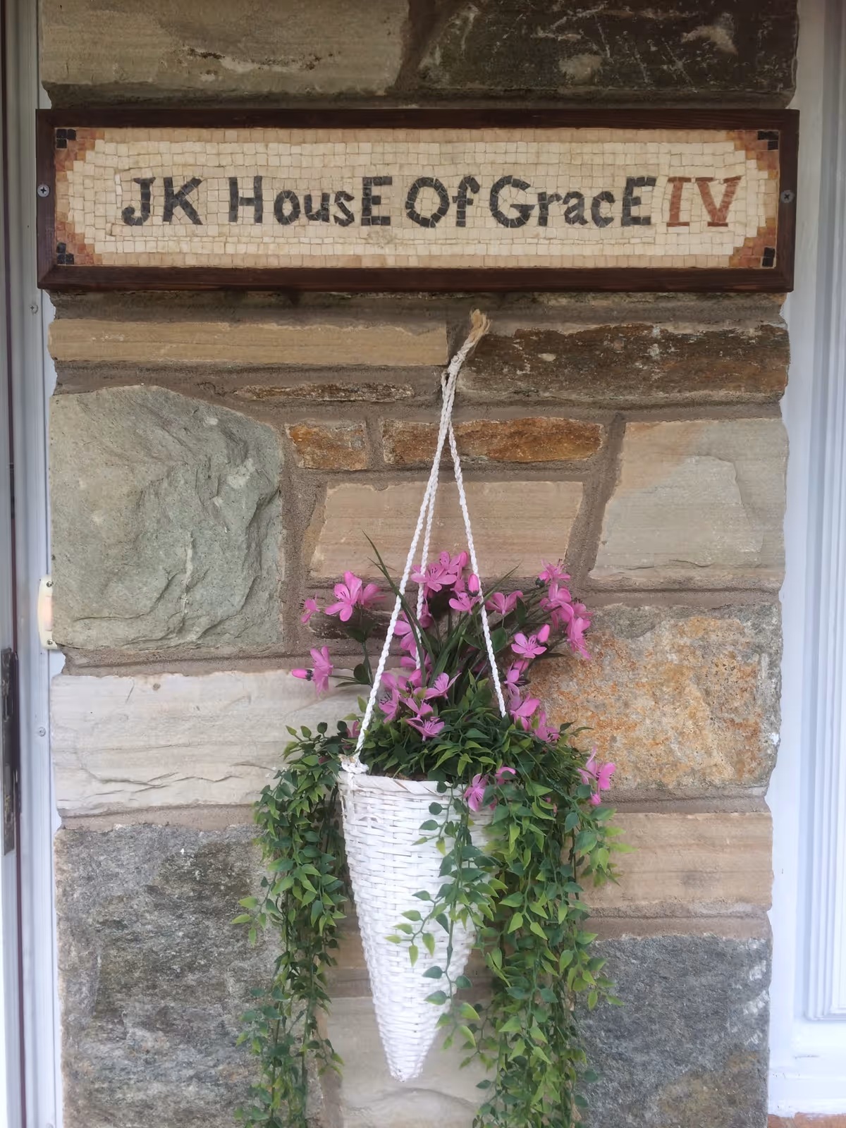 A stone wall with a mosaic sign that reads 'JK House Of Grace IV' mounted above a white hanging basket filled with pink flowers and green trailing foliage.