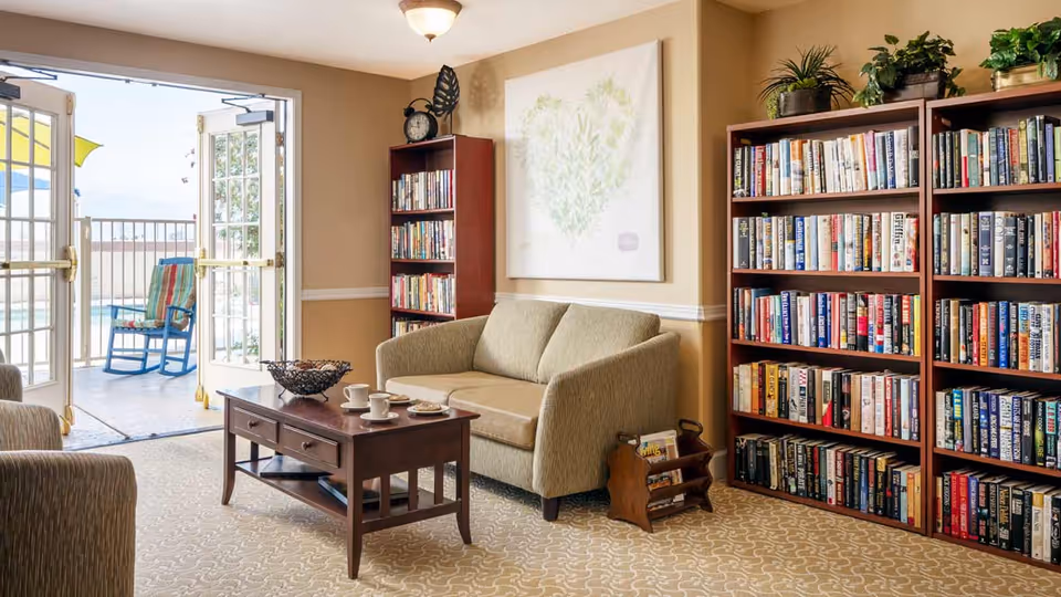 A cozy living room area in a senior living facility with a beige loveseat, two bookshelves filled with books, a wooden coffee table with cups and plates, and an open door leading to an outdoor patio with a rocking chair and umbrella.