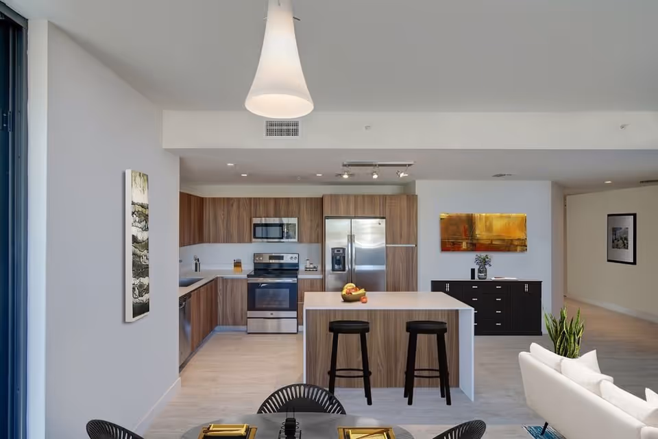 Modern kitchen and dining area with wooden cabinets, stainless steel appliances including a refrigerator, oven, and microwave. A kitchen island with two black stools is centered in the room. A white pendant light hangs above the island. In the foreground, part of a dining table with black chairs is visible. The walls are decorated with abstract artwork and a black sideboard with a vase of flowers.