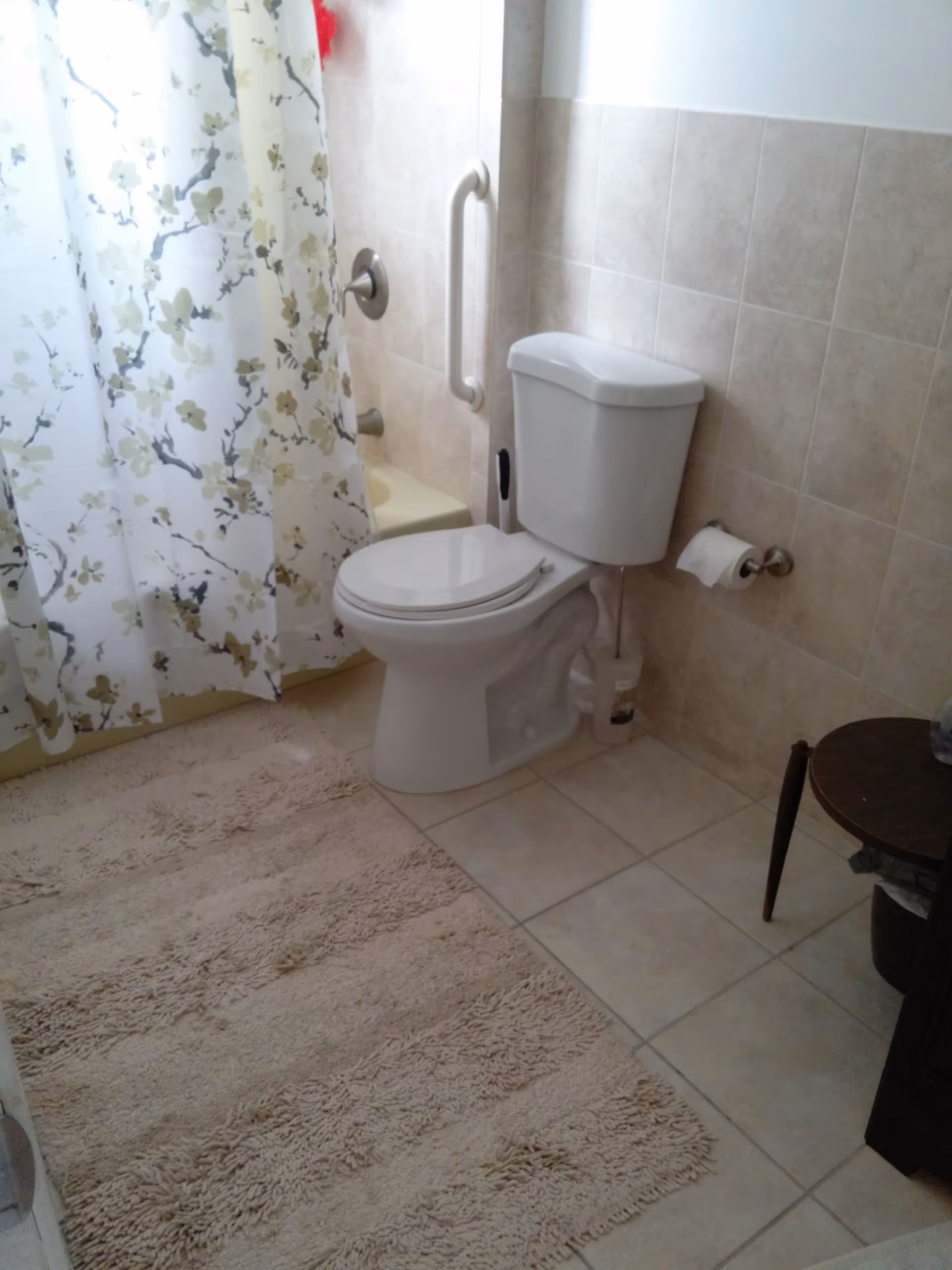 A bathroom with a white toilet, beige tiled walls and floor, a beige fluffy bath mat, a floral shower curtain, a grab bar on the wall next to the bathtub, a toilet paper holder with a roll of toilet paper, and a small round wooden side table.