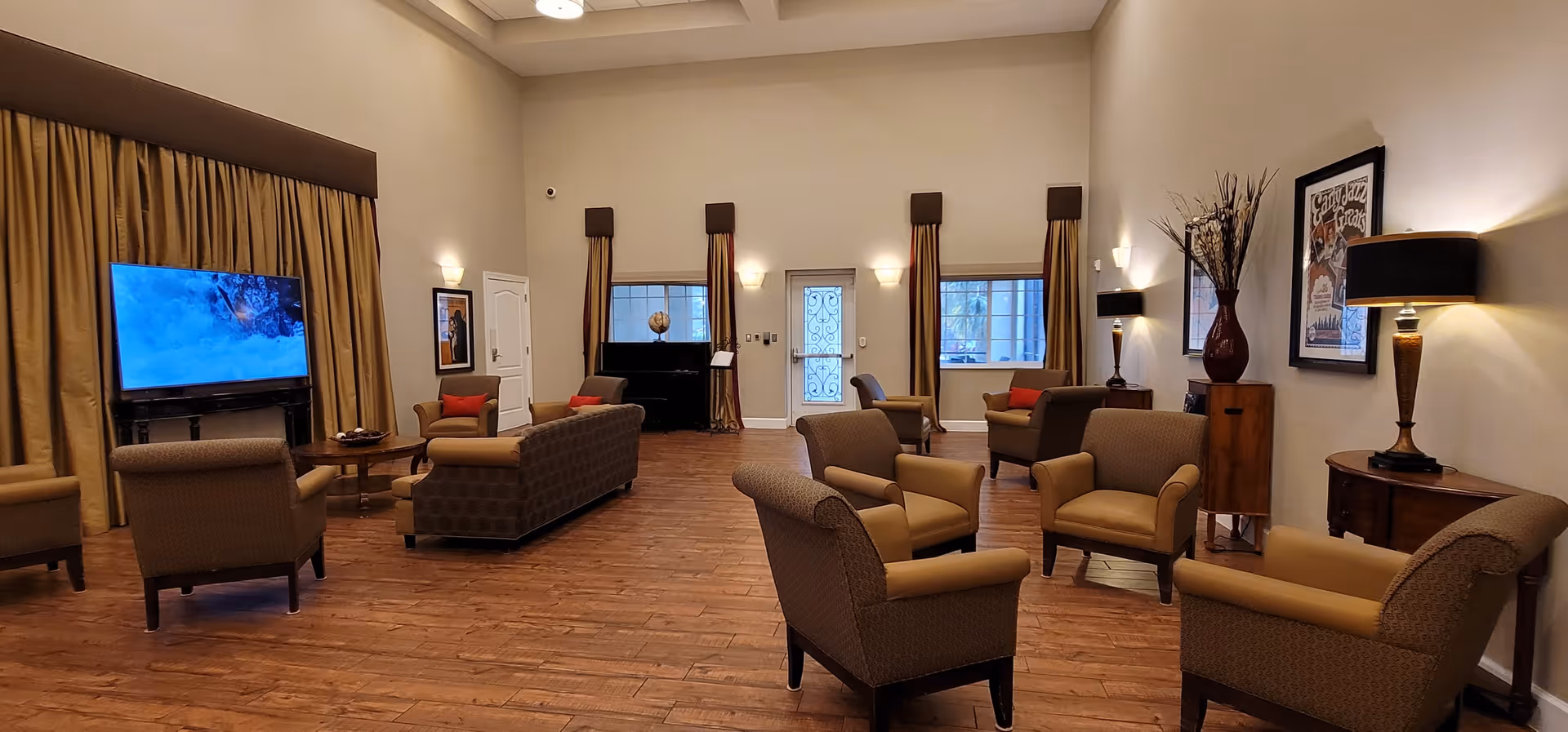 A spacious living room with wooden flooring, multiple armchairs and sofas arranged in small groups, a large flat-screen TV mounted on a stand with a nature scene displayed, tall windows with brown curtains, wall-mounted lights, decorative vases, framed artwork, and a piano in the background.