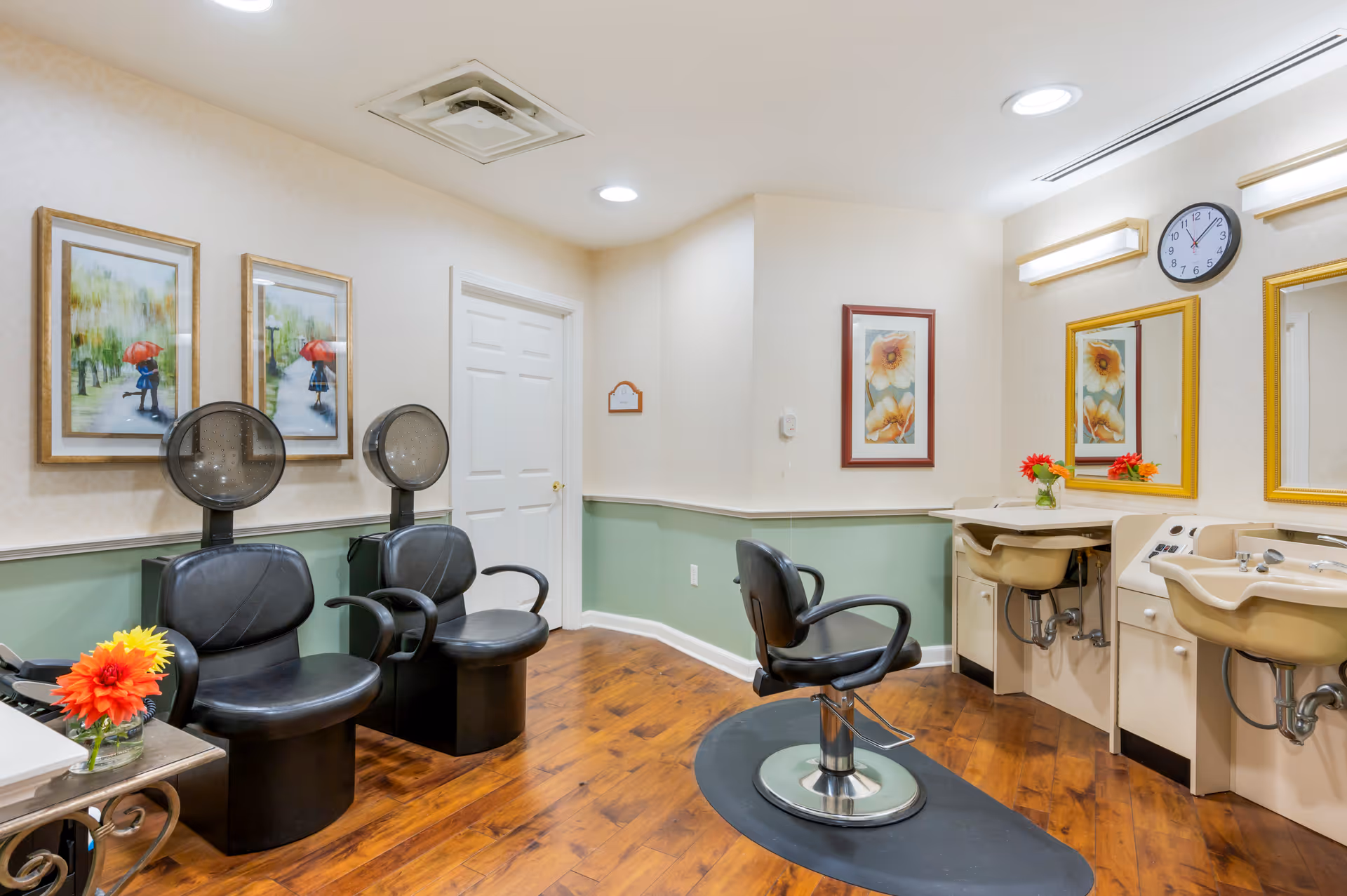 Interior of a salon room with two black hair drying chairs on the left, a single black styling chair in front of two sinks and mirrors on the right. The room has wood flooring, light green and beige walls, framed artwork, and a clock above the mirrors. A small table with a vase of orange flowers is visible on the left.