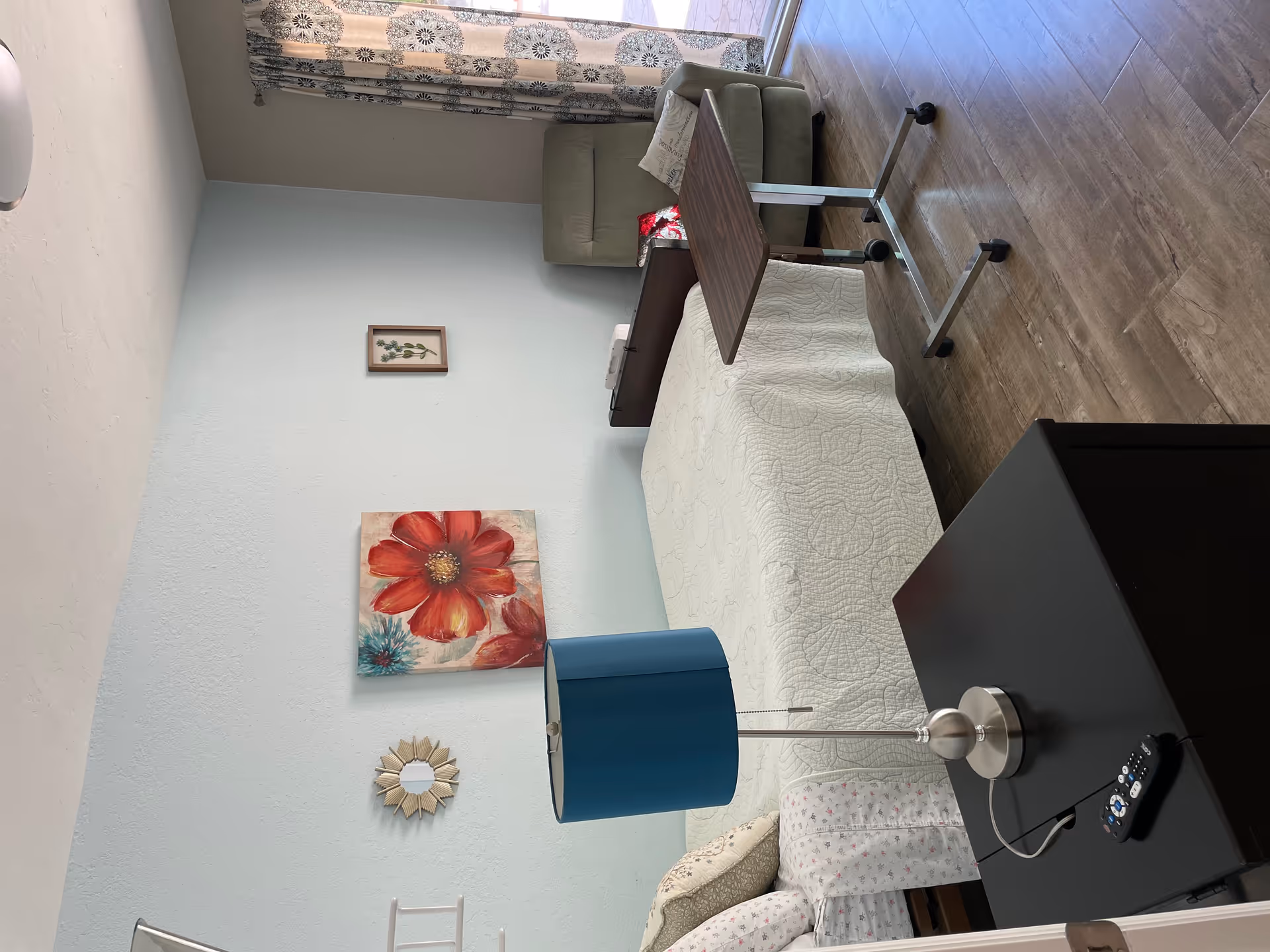 A senior living bedroom with a single bed covered in a white quilt, a black nightstand with a blue lamp and a remote control on it, a green recliner chair with pillows, a wooden overbed table on wheels, and light blue walls decorated with floral artwork and a small mirror. A sliding glass door with patterned curtains lets in natural light.