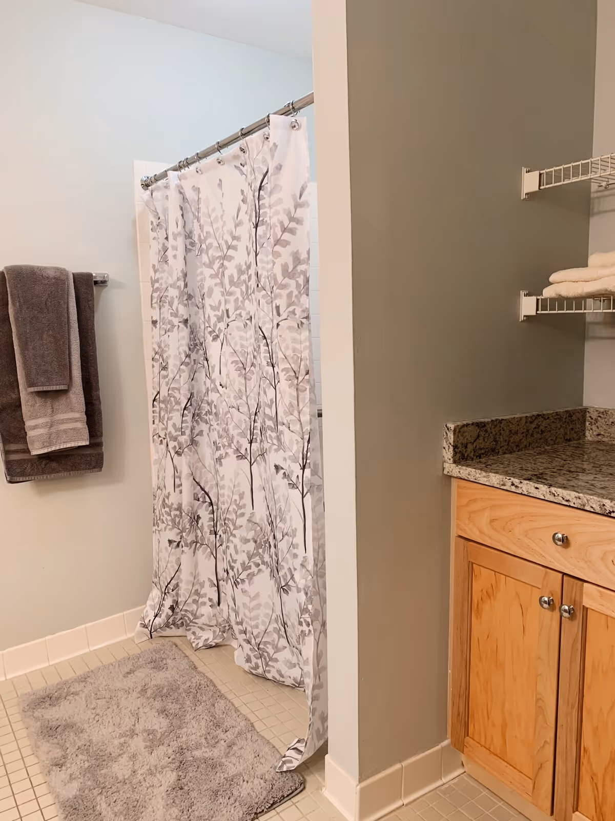 Bathroom with a shower area covered by a white curtain with gray leaf patterns, a gray bath mat on the tiled floor, two gray towels hanging on a towel rack, a wooden cabinet with a granite countertop, and wire shelves holding folded towels.