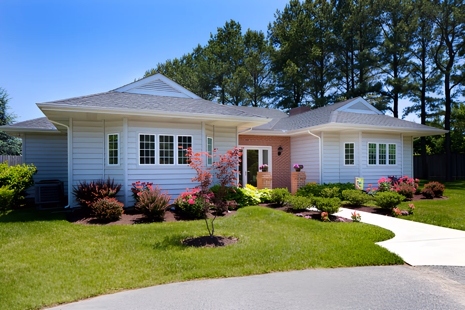 Exterior view of a single-story building with white siding and a gray roof, surrounded by a well-maintained lawn and landscaped garden with shrubs and flowers under a clear blue sky.