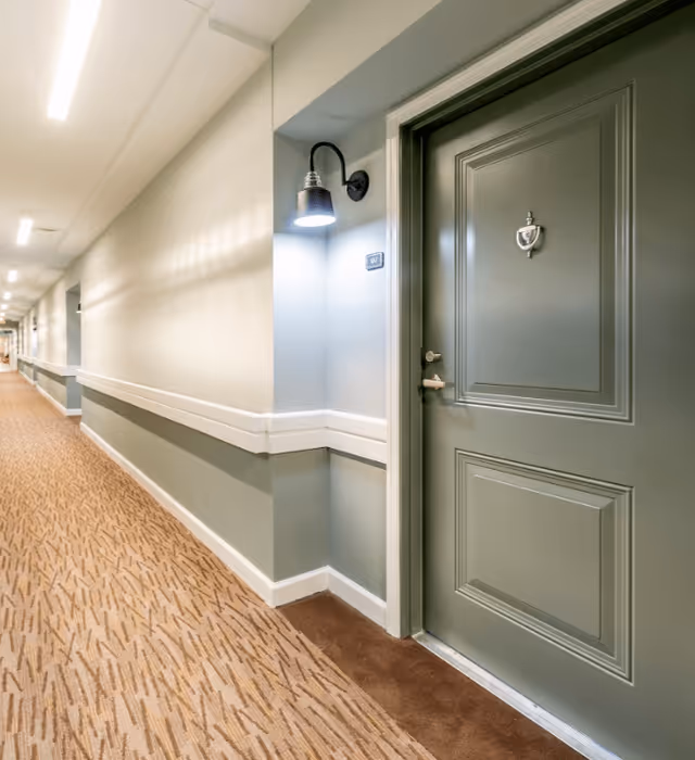 Well-lit carpeted interior hallway showing a green apartment door with a wall sconce and door knocker.