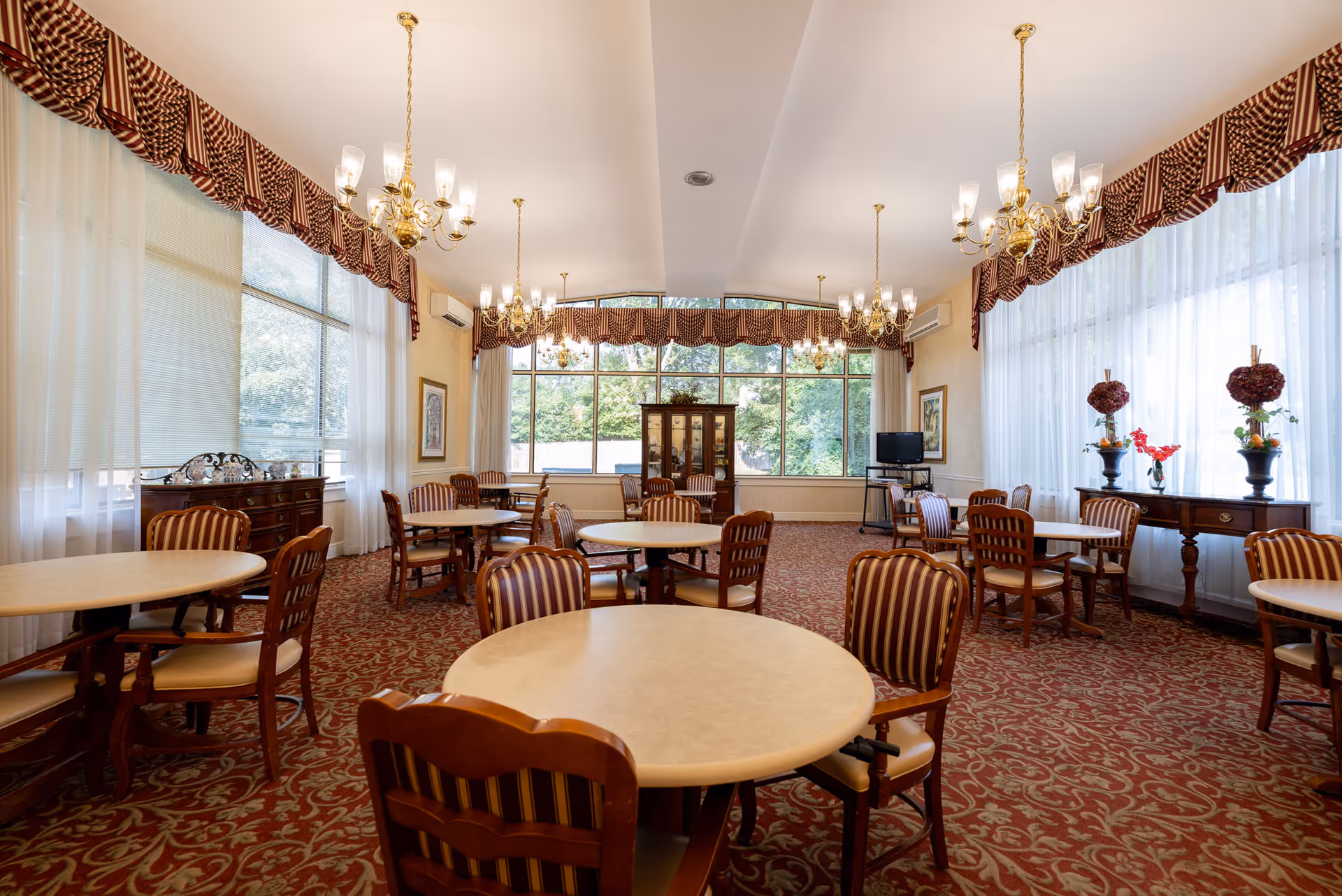 A spacious dining room with round tables and wooden chairs featuring striped upholstery. The room has large windows with sheer white curtains and patterned valances, ornate chandeliers hanging from the ceiling, and a carpet with a red and beige floral pattern. There are decorative vases with flowers on side tables and a cabinet against the far wall.