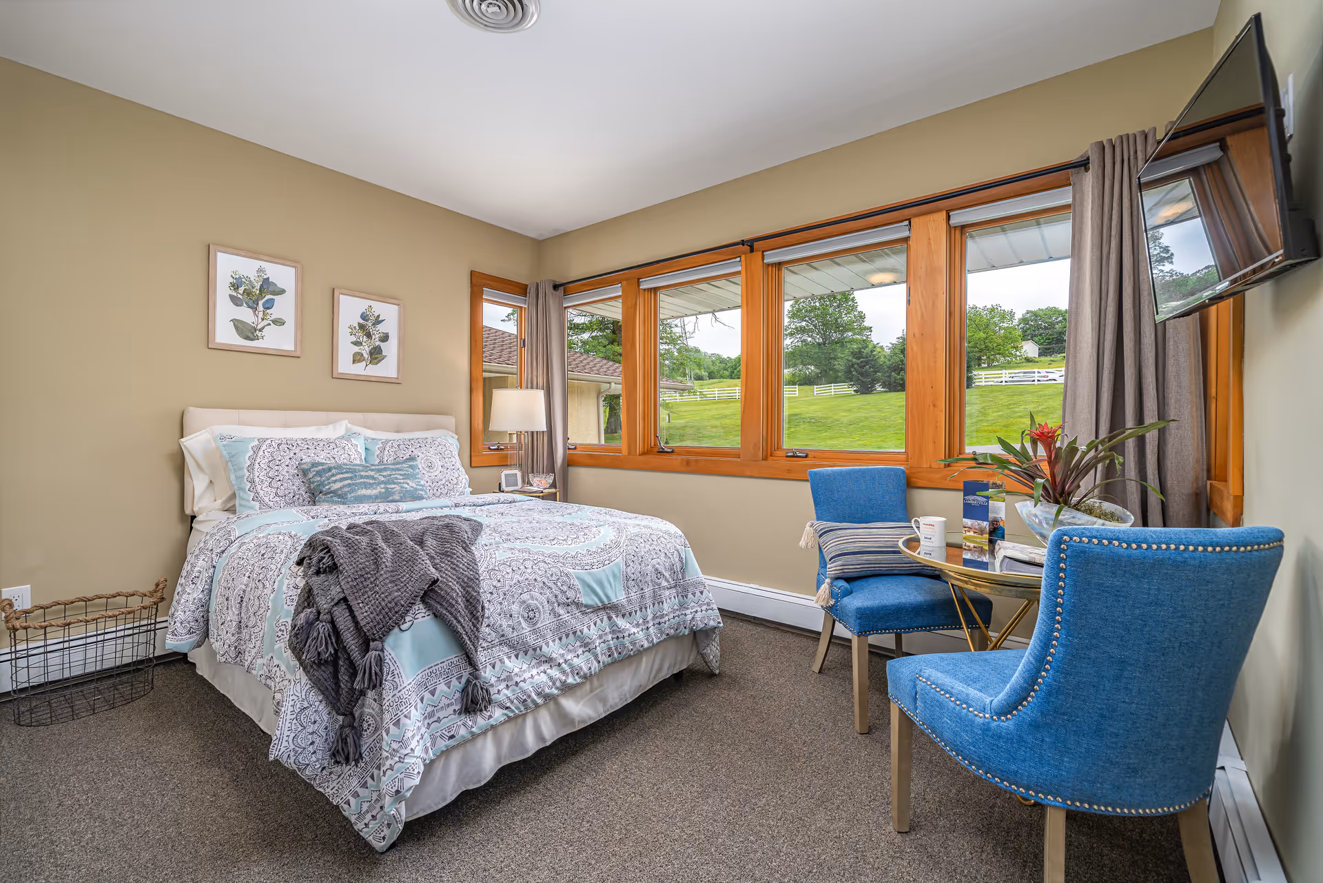 A cozy bedroom featuring a large bed with decorative bedding, two blue chairs, a small table with a plant and a mug, and large windows offering a view of greenery outside.