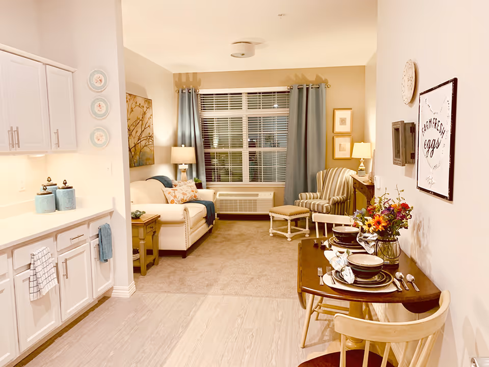Open-plan interior showing a living area with a sofa and armchair by a window, a small set dining table on the right, and a kitchen counter on the left.