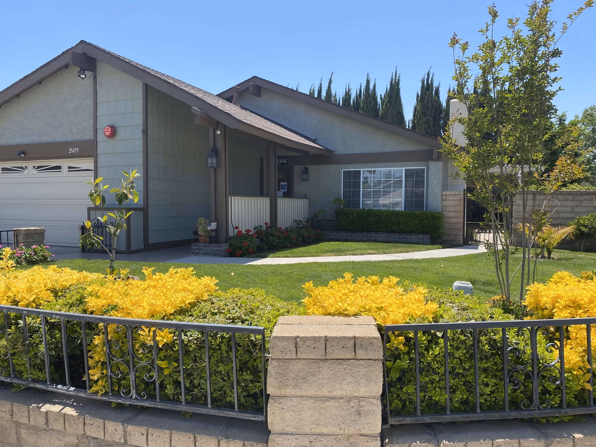 Single-story residential building with a well-maintained front yard featuring green grass, yellow and green shrubs, a small tree, and a concrete pathway leading to the entrance. The house has a garage on the left side and a covered porch area with some potted plants and flowers near the door. The sky is clear and blue.