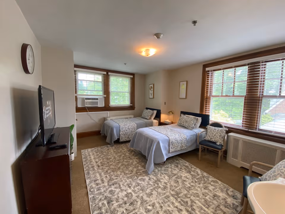 A bright bedroom with two single beds covered in light blue bedding and patterned runners. Each bed has a decorative pillow and a nightstand with a lamp between them. The room has large windows with wooden blinds letting in natural light, a patterned area rug on the floor, a wall clock, a TV on a dark wooden dresser, and two chairs with cushions. There is also a small sink visible in the corner.