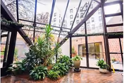 Glass-roofed indoor atrium with large potted plants, brick flooring, and glass doors/windows opening to a multi-story building.