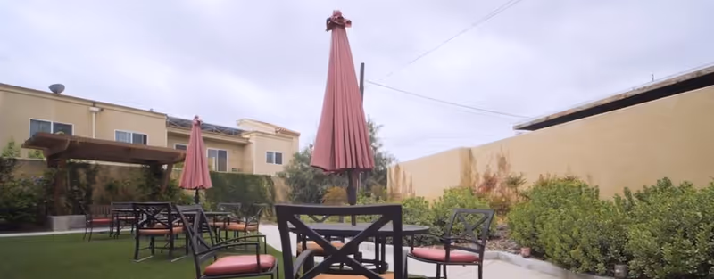 Outdoor seating area in a senior living facility with multiple black metal tables and chairs with red cushions, two closed red umbrellas, surrounded by green bushes and plants, with a beige building and a pergola in the background under a cloudy sky.