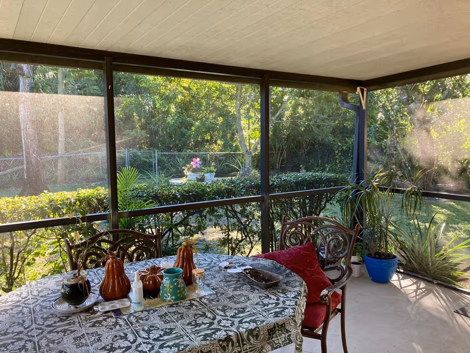 A screened-in porch area with a table covered by a patterned tablecloth. On the table are decorative items including ceramic pumpkins, a green pitcher, a small bottle, and a plant. Two ornate chairs with red cushions are positioned around the table. Outside the screened porch, there is greenery including bushes, trees, and potted plants.
