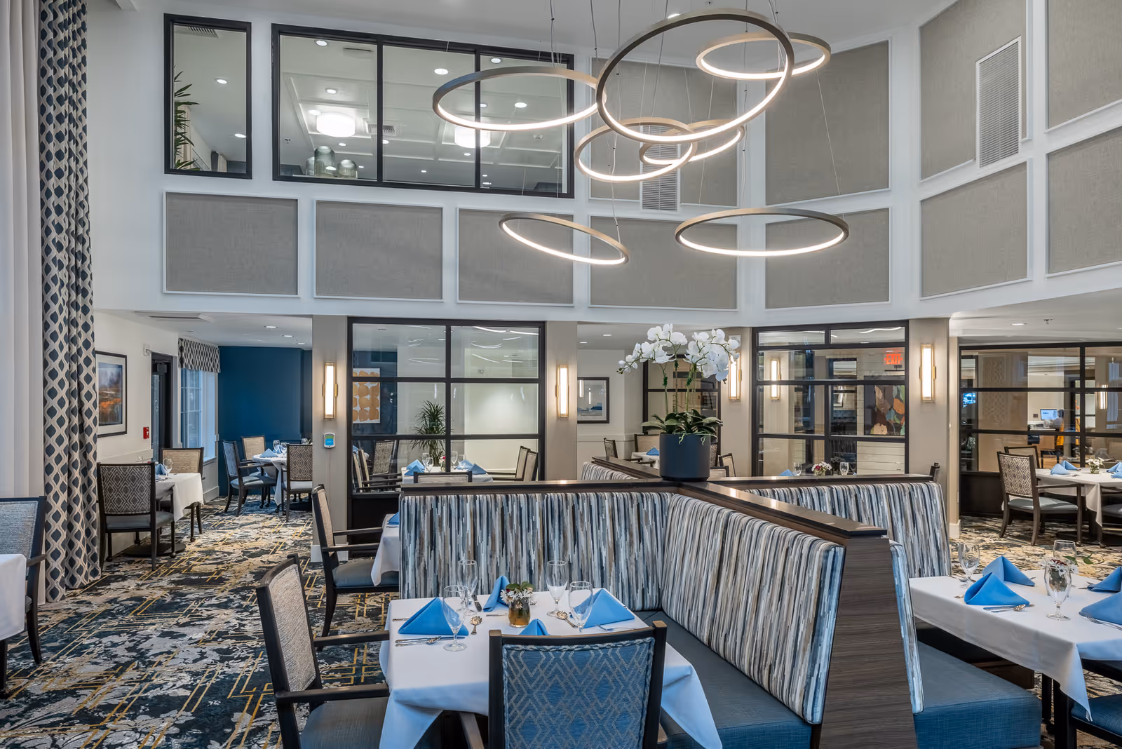 A bright and spacious dining room in a senior living facility with modern circular pendant lights hanging from a high ceiling. The room features tables set with white tablecloths, blue folded napkins, glassware, and small floral centerpieces. Upholstered booth seating with striped fabric divides the space, and additional chairs with patterned upholstery surround the tables. Large windows and glass partitions separate this area from other rooms, and decorative wall panels and curtains add to the elegant ambiance.