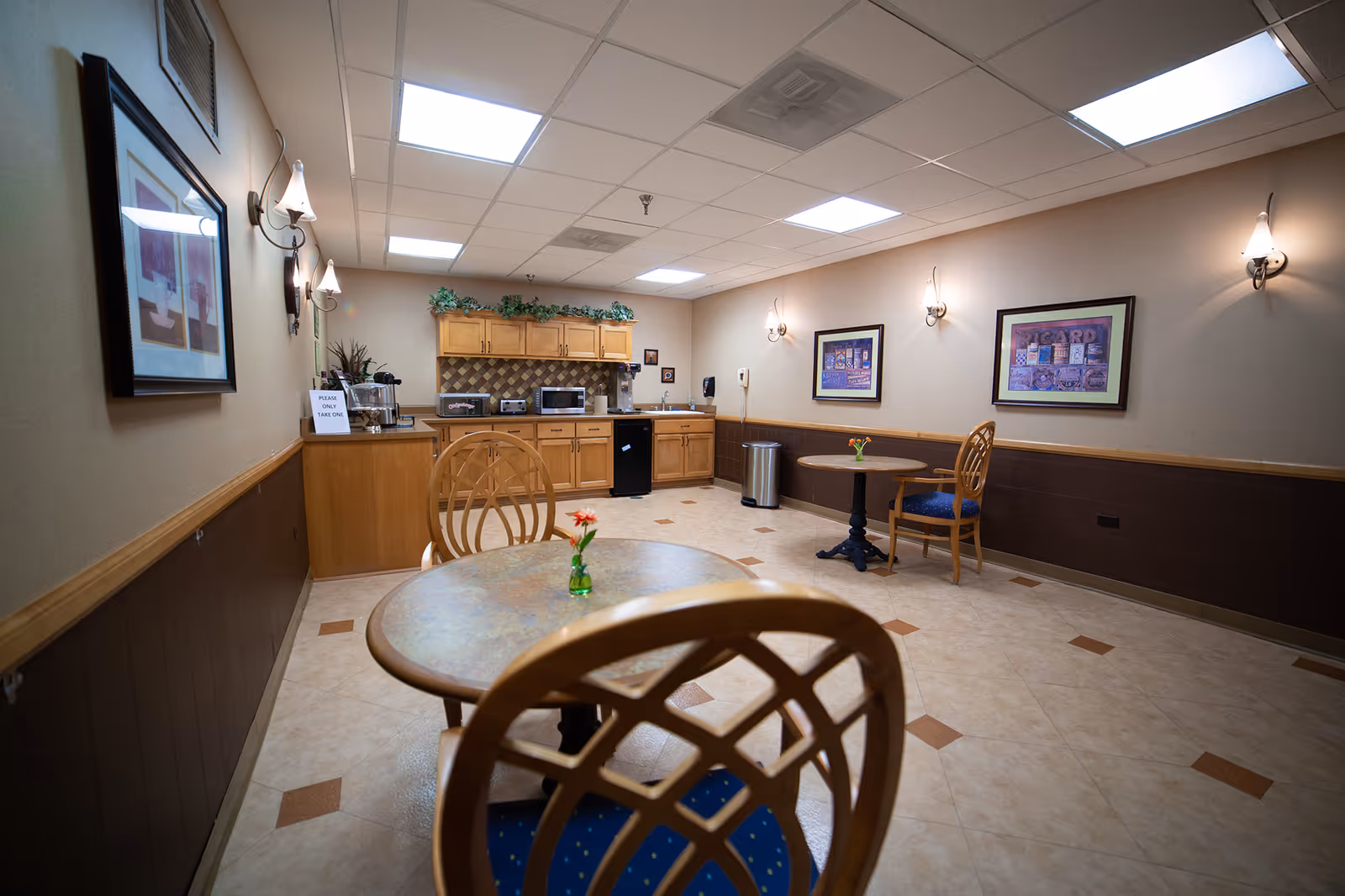 Small communal dining area with round tables and chairs and a kitchenette with cabinets and appliances along the back wall.