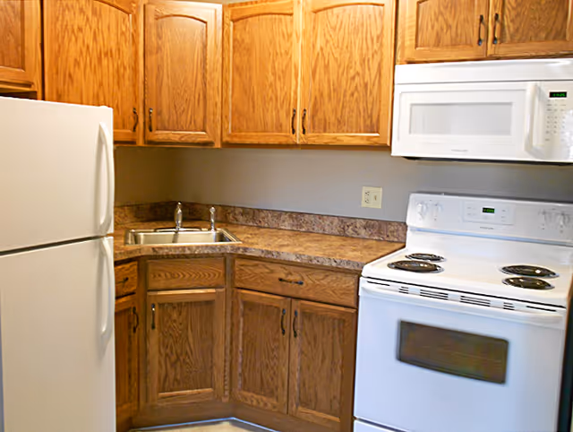 A small kitchen area with wooden cabinets, a white refrigerator on the left, a stainless steel sink in the corner, and a white electric stove with an overhead microwave on the right.