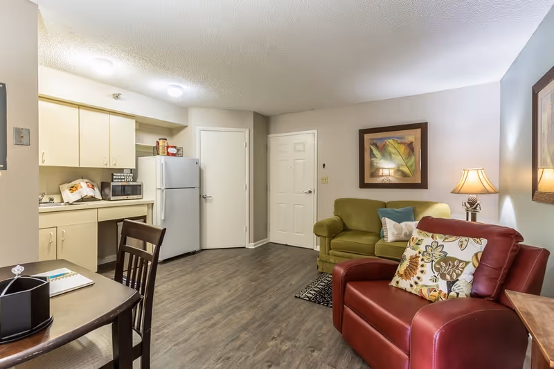 A cozy living area with a small kitchen in the background. The kitchen has cream-colored cabinets, a white refrigerator, and a microwave. In the living area, there is a green loveseat with decorative pillows, a red armchair with a floral pillow, a wooden side table with a lamp, and a framed artwork on the wall. The floor is wood laminate, and the walls are painted in neutral tones.