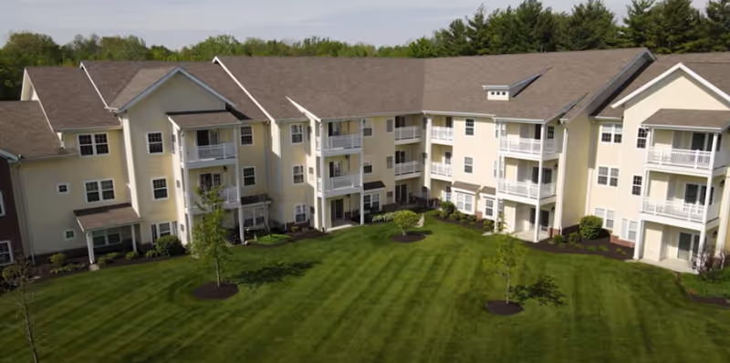 A large, three-story residential building with beige siding and multiple balconies overlooking a well-maintained green lawn with small trees and landscaped areas.