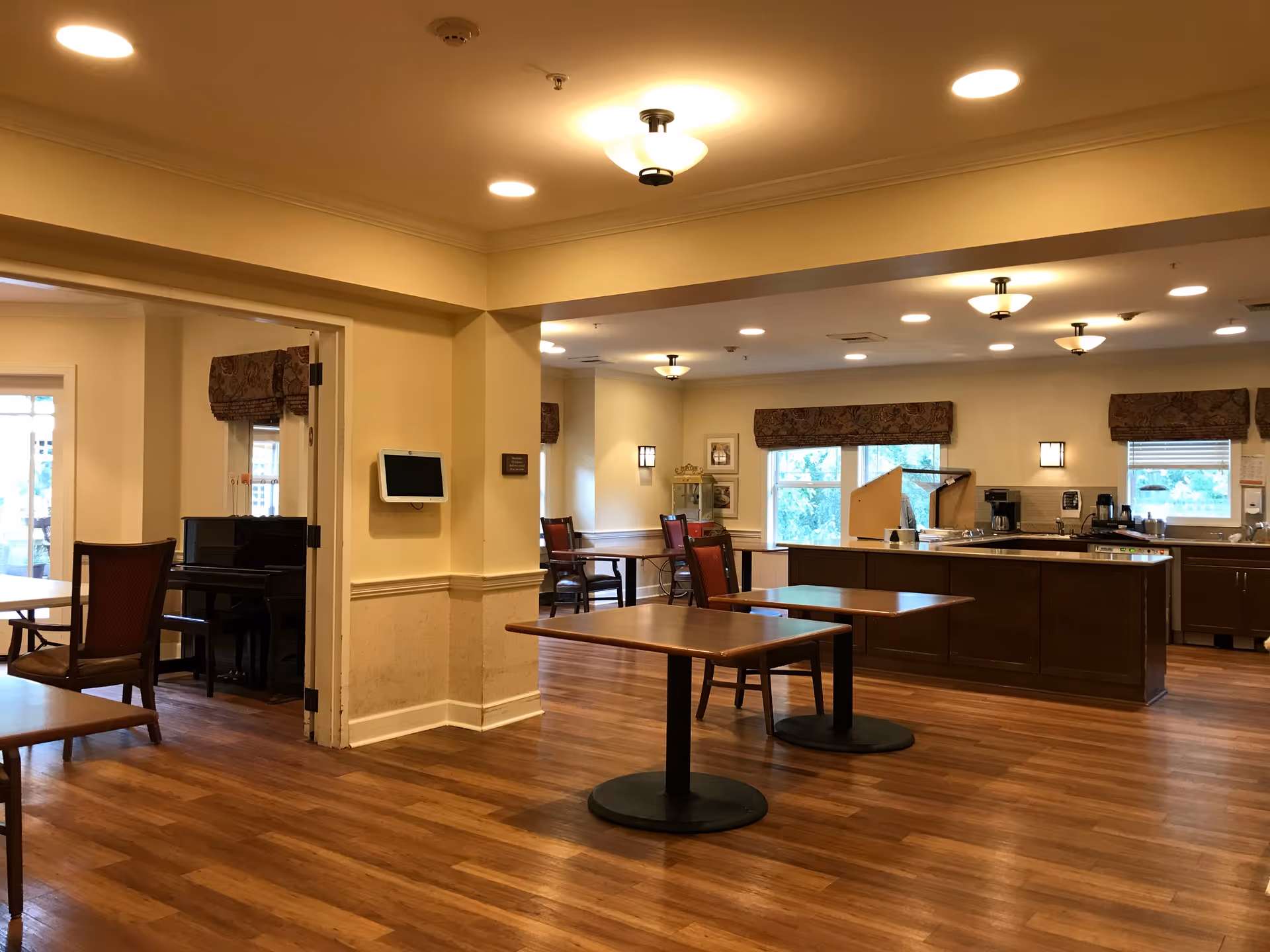 Spacious senior living dining/common area with wooden floors, empty tables and chairs and a kitchen island under warm ceiling lights.