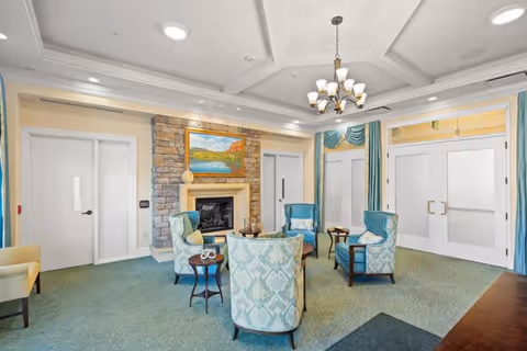 A cozy common area with four patterned armchairs arranged around a small round table in front of a stone fireplace with a landscape painting above it. The room has light-colored walls, teal carpet, and large windows with teal and yellow curtains. A chandelier hangs from the ceiling, and there are white double doors on one side.