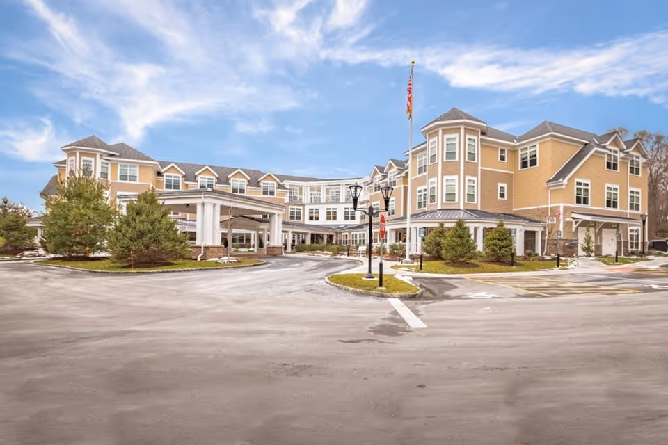 Exterior view of a large, multi-story senior living facility building with beige siding and white trim. The building has multiple windows, a covered entrance, and an American flag on a flagpole in front. There are small trees and landscaped areas around the driveway and parking lot under a partly cloudy sky.