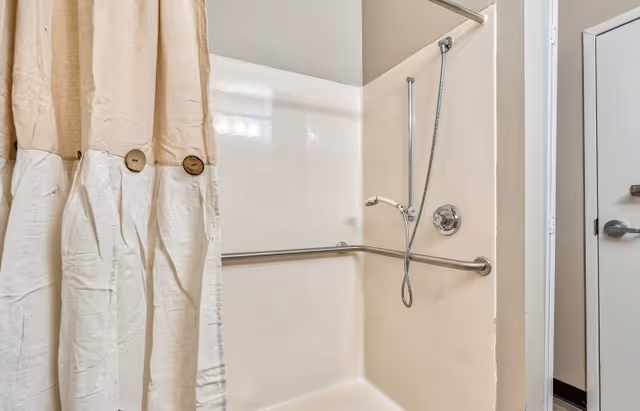 A shower area with a beige and white shower curtain featuring wooden buttons, a handheld showerhead attached to a flexible hose, and a horizontal grab bar on the wall. The shower is enclosed with light-colored walls and a door with a metal handle is visible to the right.