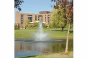Multi-story brick building set behind a pond with a central fountain and surrounding trees on a grassy lawn.