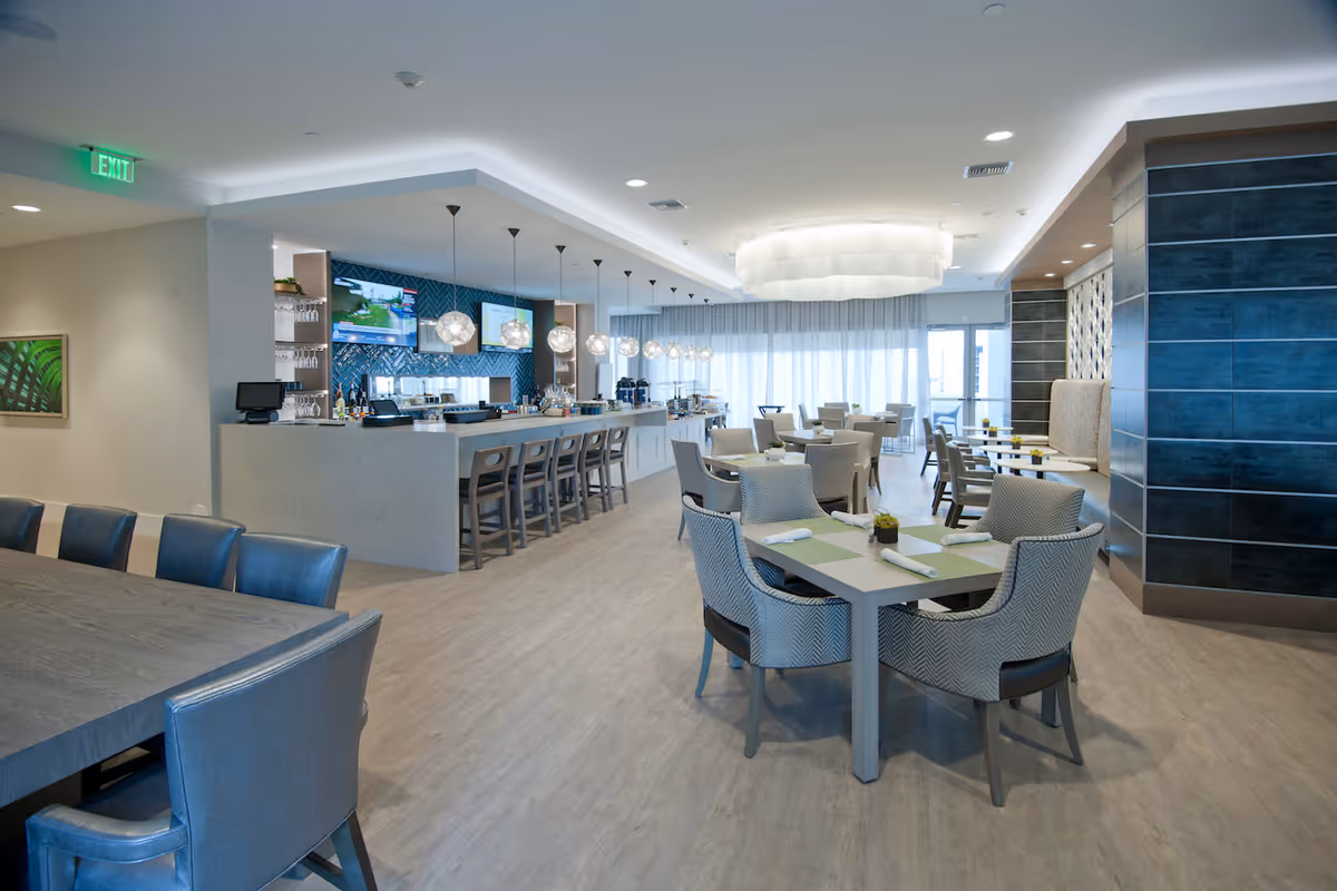 Modern dining room with multiple tables and chairs, a long bar with stools, pendant lighting, and large windows.