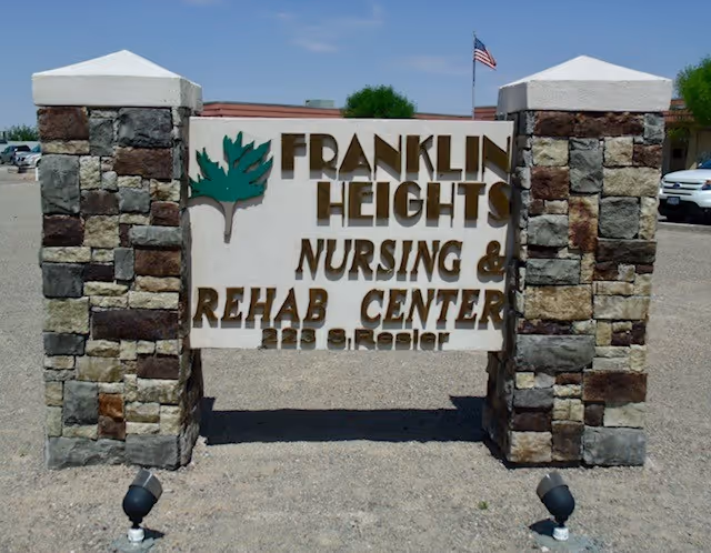 Stone and concrete sign for Franklin Heights Nursing & Rehab Center with an American flag in the background and parked cars on a gravel surface.