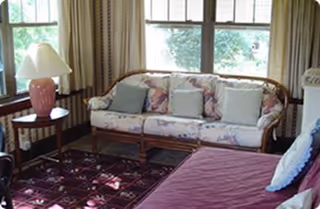 A cozy living room area with a floral-patterned sofa adorned with several cushions, a wooden side table with a pink lamp, large windows with curtains allowing natural light, and a purple upholstered chair in the foreground.