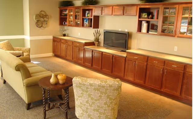 Common area with a green sofa and patterned armchair facing a TV on long wooden built-in cabinets and shelving.