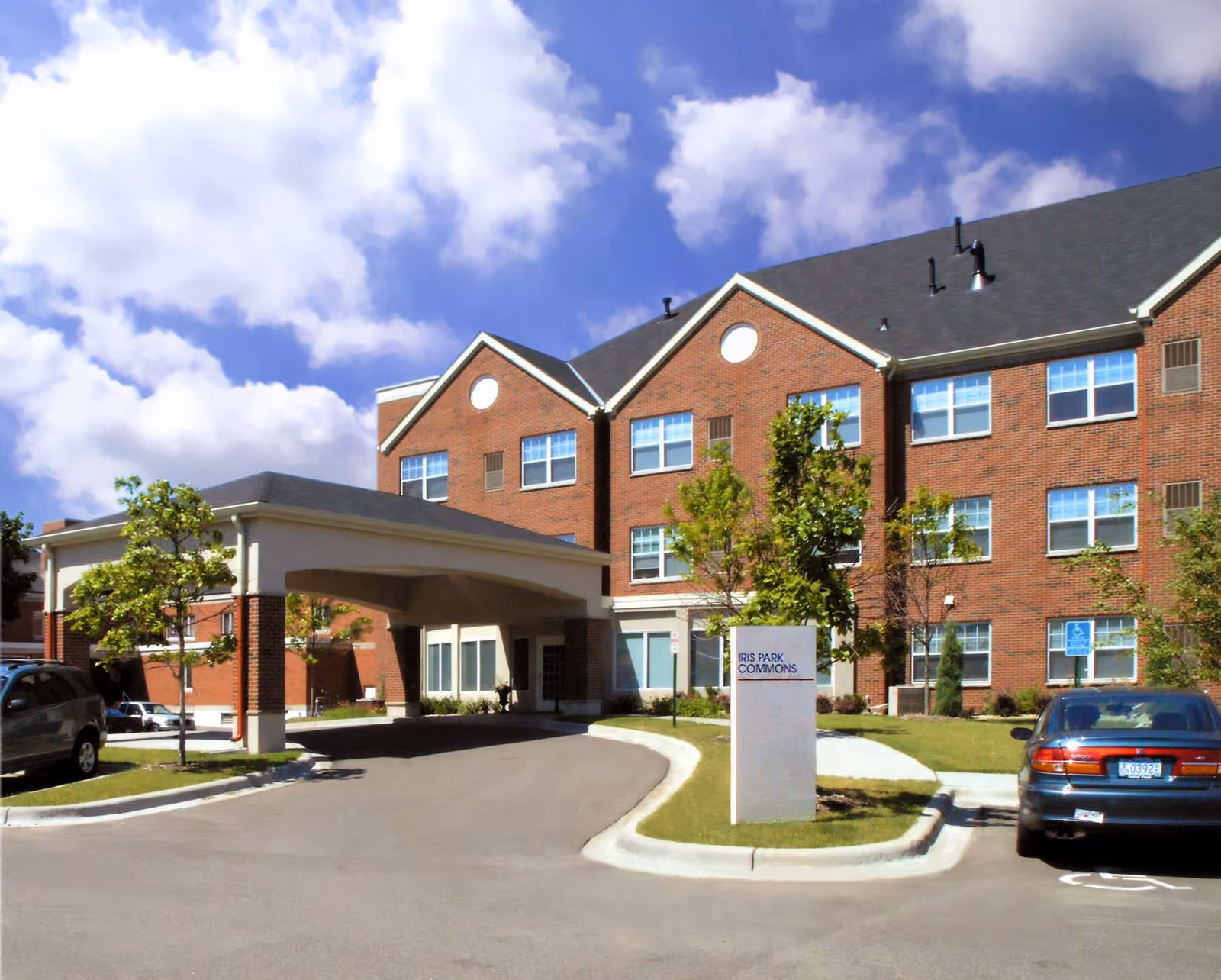 Exterior view of Iris Park Commons Assisted Living by Episcopal Homes, showing a three-story brick building with multiple windows, a covered entrance driveway, several small trees, parked cars, and a sign with the facility name.