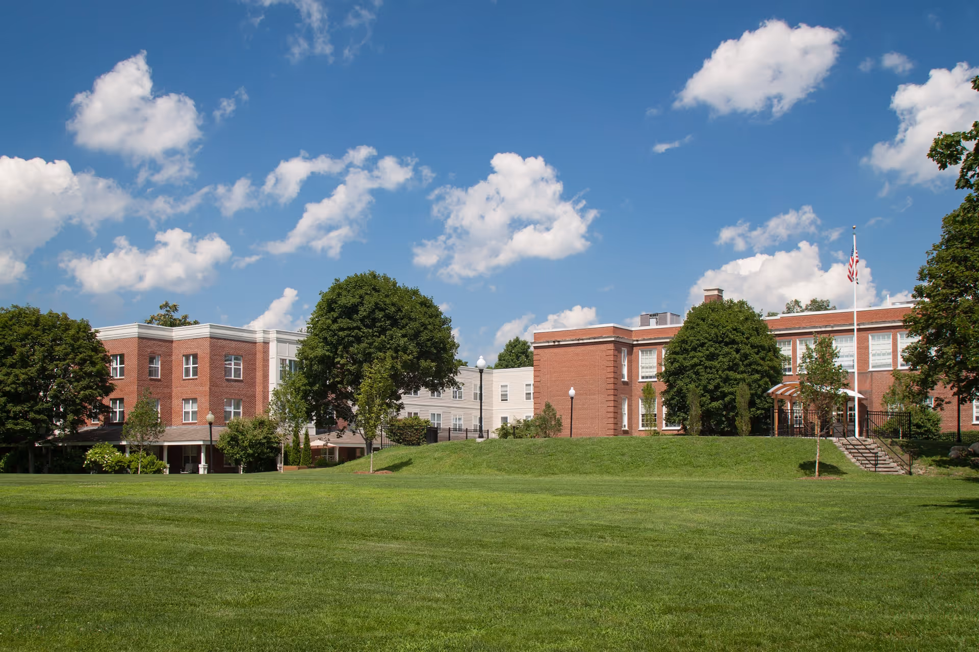 A large, well-maintained green lawn in front of a multi-story brick and white building under a blue sky with scattered clouds. The building is surrounded by trees and has an American flag on a flagpole near the entrance.