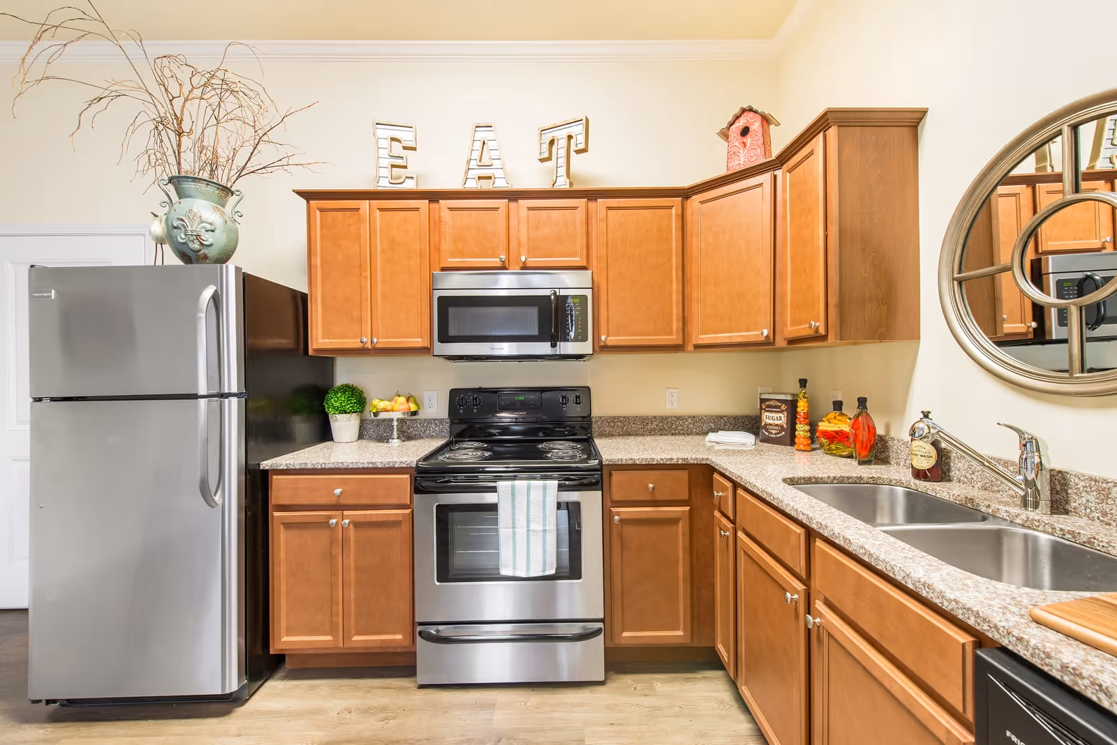 A modern kitchen with wooden cabinets, a stainless steel refrigerator, stove, and microwave. The countertop has a double sink, decorative items, and a towel hanging on the oven handle. Above the cabinets, large letters spell out 'EAT'. A round mirror is mounted on the wall to the right.