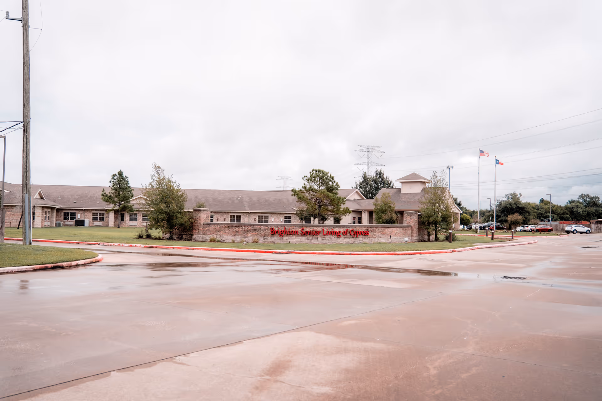 Exterior view of Brighton Senior Living facility showing a single-story brick building with a sign in front that reads 'Brighton Senior Living of Cypress'. The scene includes a wet driveway, some trees, and two flagpoles with the American and Texas flags.