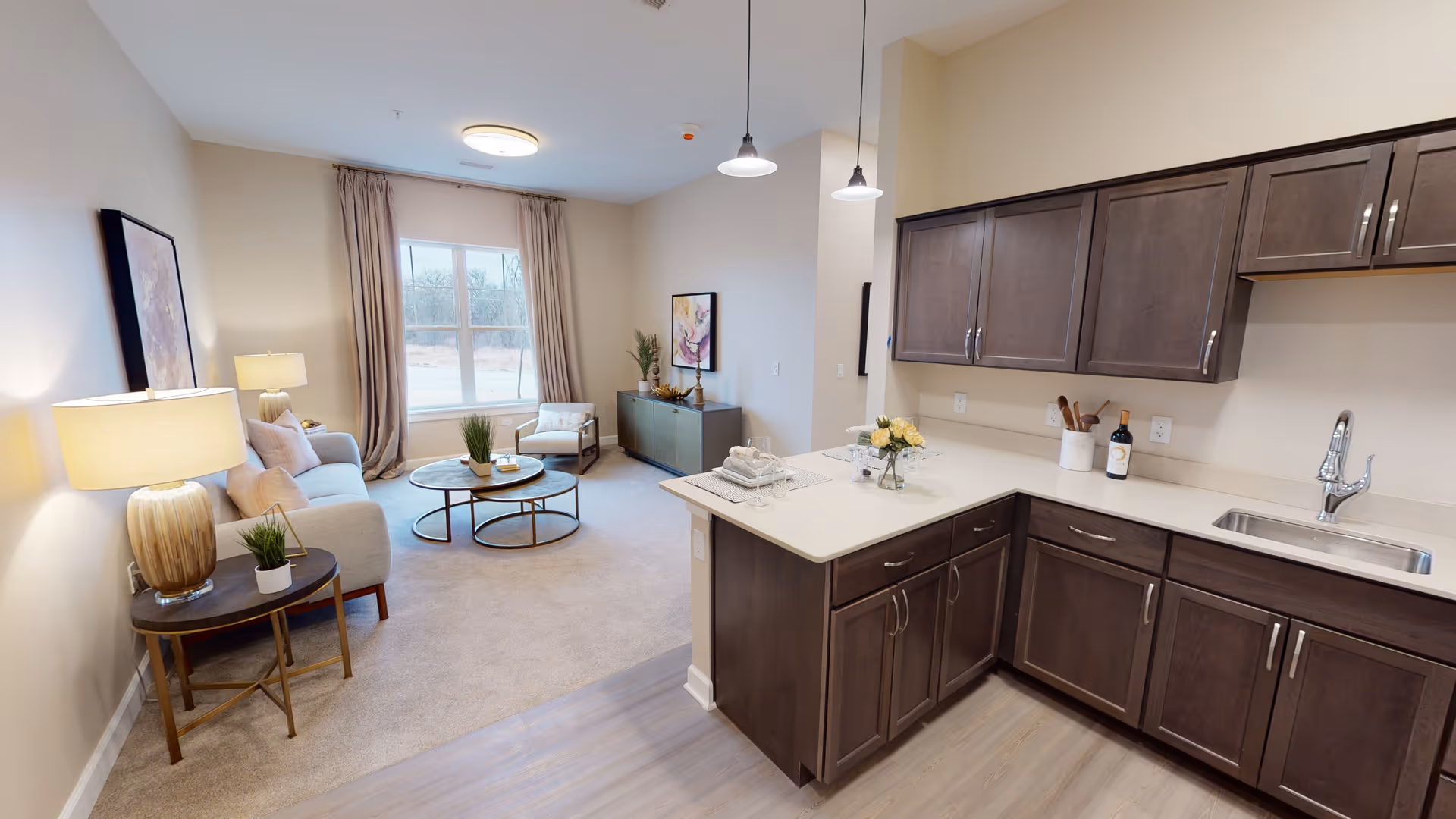 A bright and modern senior living apartment interior featuring a kitchen with dark wood cabinets and a white countertop on the right, and a cozy living room area on the left with a sofa, armchair, round coffee table, side table with a lamp, and a large window with beige curtains letting in natural light.