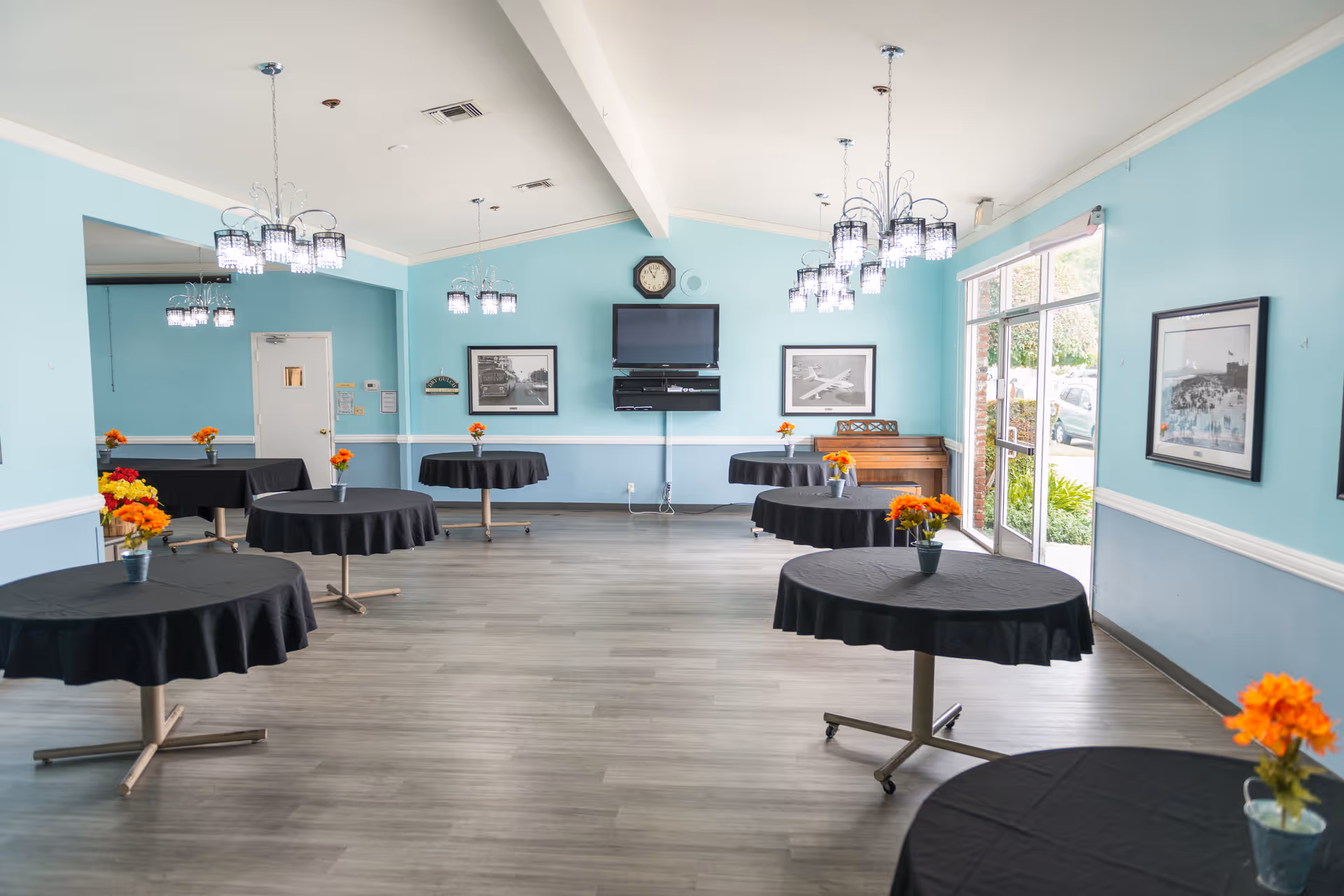 A bright dining room with light blue walls and several round tables covered with black tablecloths, each decorated with a small vase of orange flowers. The room features modern chandeliers, a wall-mounted TV, framed pictures, a piano, and large glass doors letting in natural light.