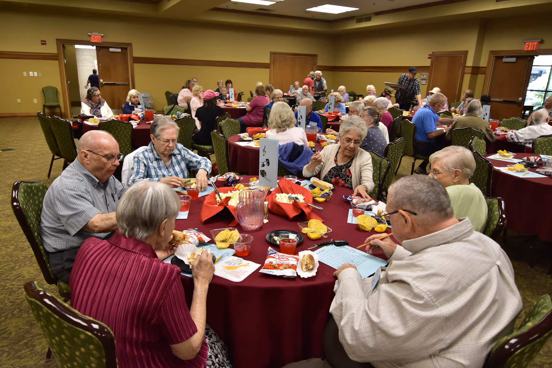 A group of elderly people sitting around multiple round tables with red tablecloths in a large dining room, eating and socializing. The room has green patterned chairs, beige walls, and exit doors in the background.