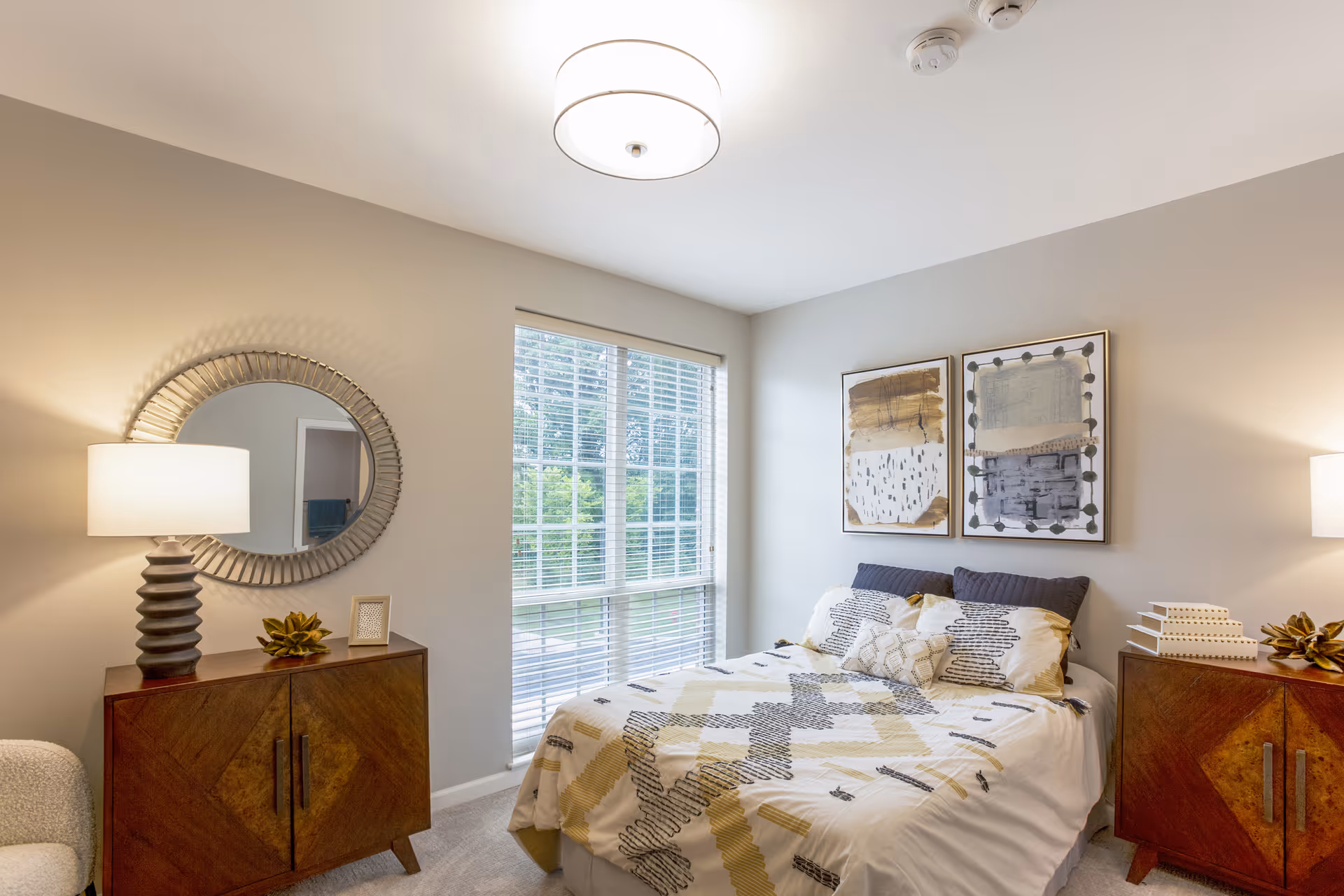 A cozy bedroom with a bed covered in a patterned comforter and multiple pillows. On either side of the bed are wooden nightstands with table lamps and decorative items. A round mirror hangs on the wall above one nightstand, and two abstract paintings are displayed above the bed. A large window with white blinds lets in natural light.