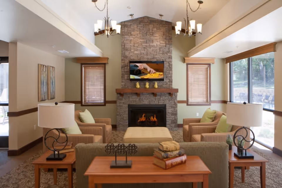 A cozy living room with a stone fireplace at the center, a flat-screen TV mounted above it displaying a cooking scene. The room has two windows with wooden blinds on either side of the fireplace, two armchairs with green cushions, a beige ottoman, and a sofa with a wooden console table behind it. Two table lamps with white shades are on side tables next to the armchairs. The room is well-lit with natural light from large windows on the right side and two chandeliers hanging from the ceiling.
