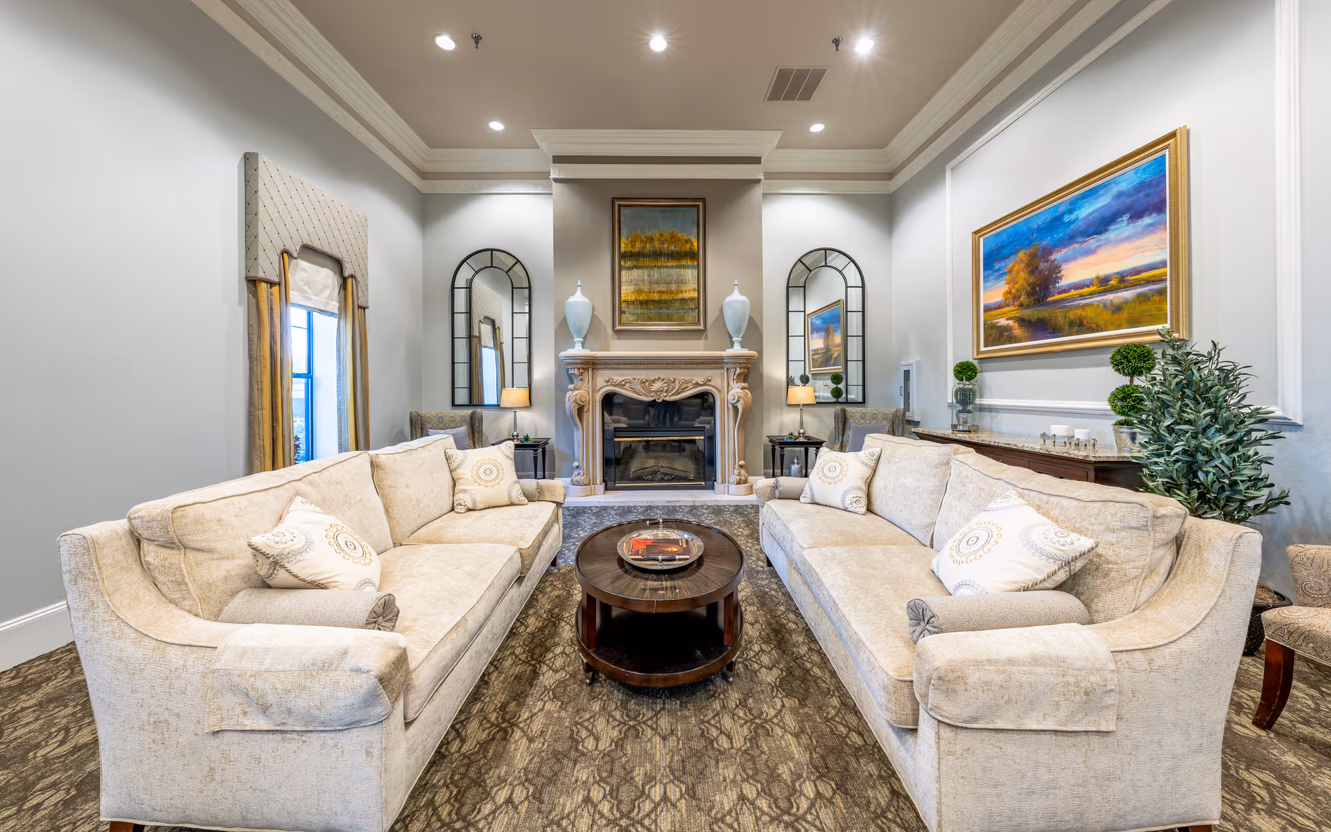 A cozy living room with two beige sofas facing each other, a round wooden coffee table in the center, a decorative fireplace with vases and a painting above it, two tall mirrors on either side of the fireplace, and a large colorful landscape painting on the right wall. The room is well-lit with recessed ceiling lights and has a patterned carpet and a potted plant near the right sofa.