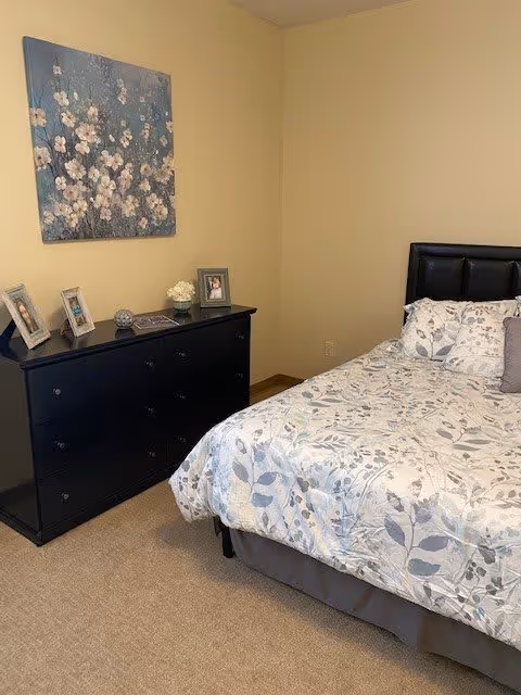 A bedroom with a bed featuring a floral patterned comforter and a black cushioned headboard. Next to the bed is a black dresser with framed photos and decorative items on top. A floral painting hangs on the beige wall above the dresser.