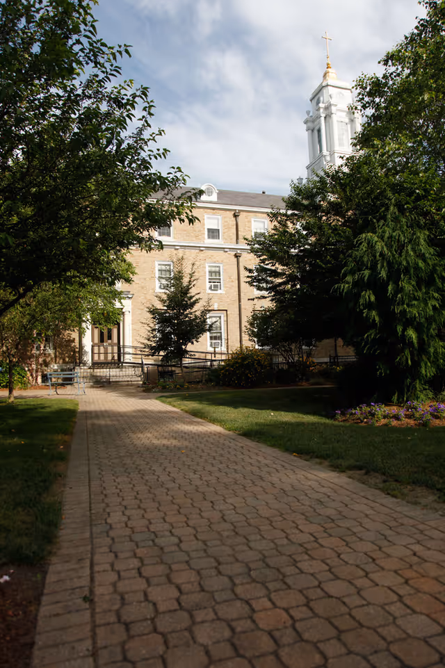 A paved walkway leading through a garden area with trees and bushes towards a multi-story brick building with white-trimmed windows and a white steeple topped with a gold cross under a partly cloudy sky.