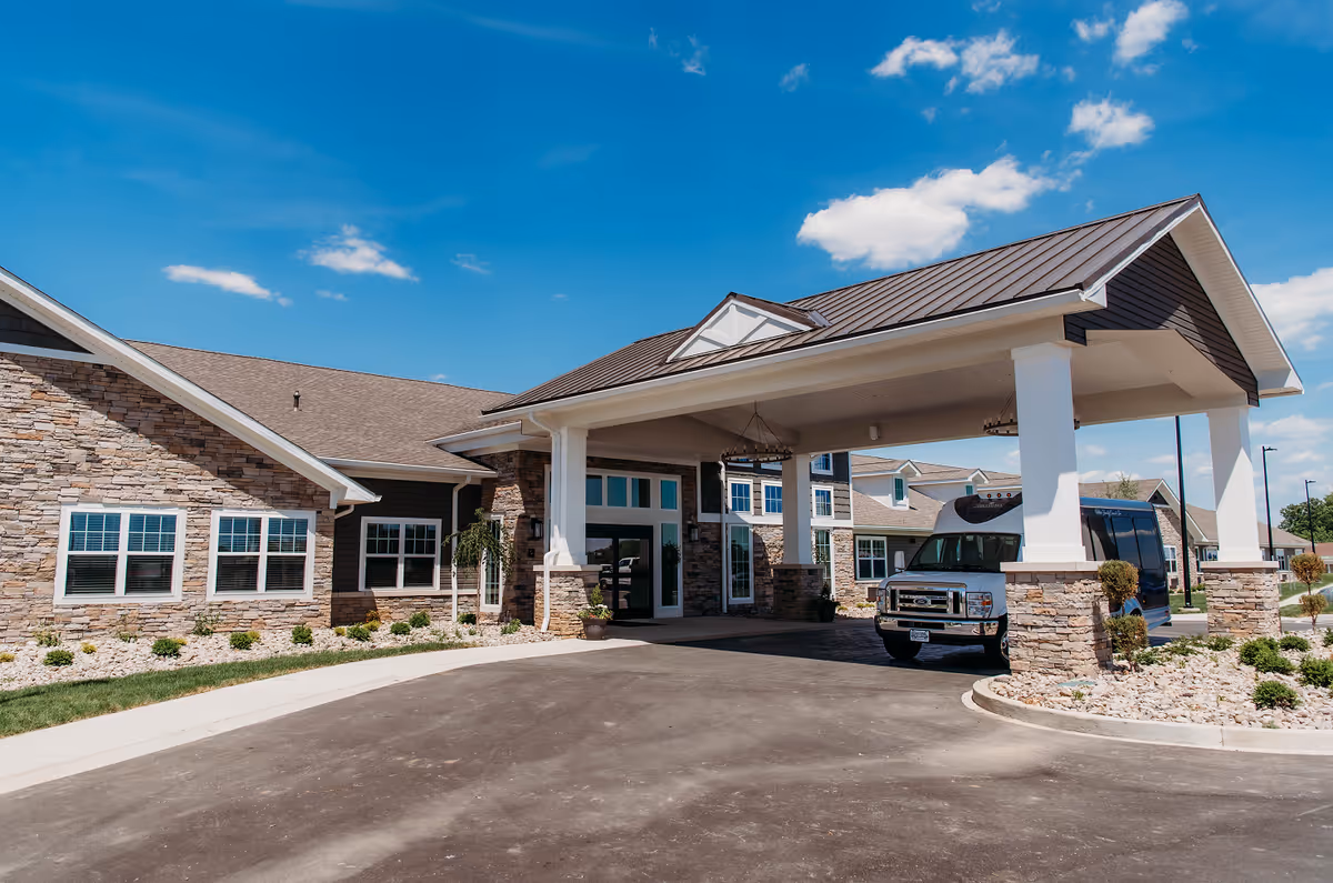 Front exterior of a senior living facility showing a covered entrance with stone facade and a parked van.